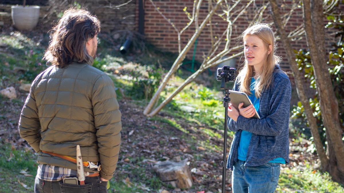 A student interviewing a person on camera. Both are standing outside.